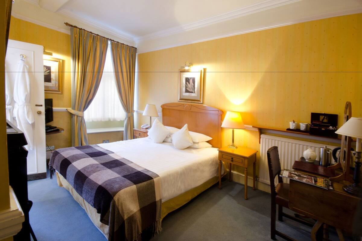 Une chambre d’hôtel comprenant un lit, des tables de chevet, un bureau, une chaise, un porte-manteau et une fenêtre avec des rideaux.