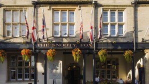 Un edificio con el letrero "The Snooty Fox" y varias banderas. Se ven cestas de flores colgantes y un cartel.