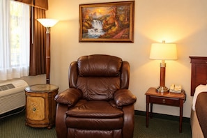 A hotel room with a leather armchair, a painting of a waterfall, a lamp, a bedside table with a phone, and a radiator. - O'cairns Inn & Suites (Lompoc)