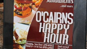 A menu board advertising O'CAIRNS HAPPY HOUR with tacos, burgers, and sandwiches.