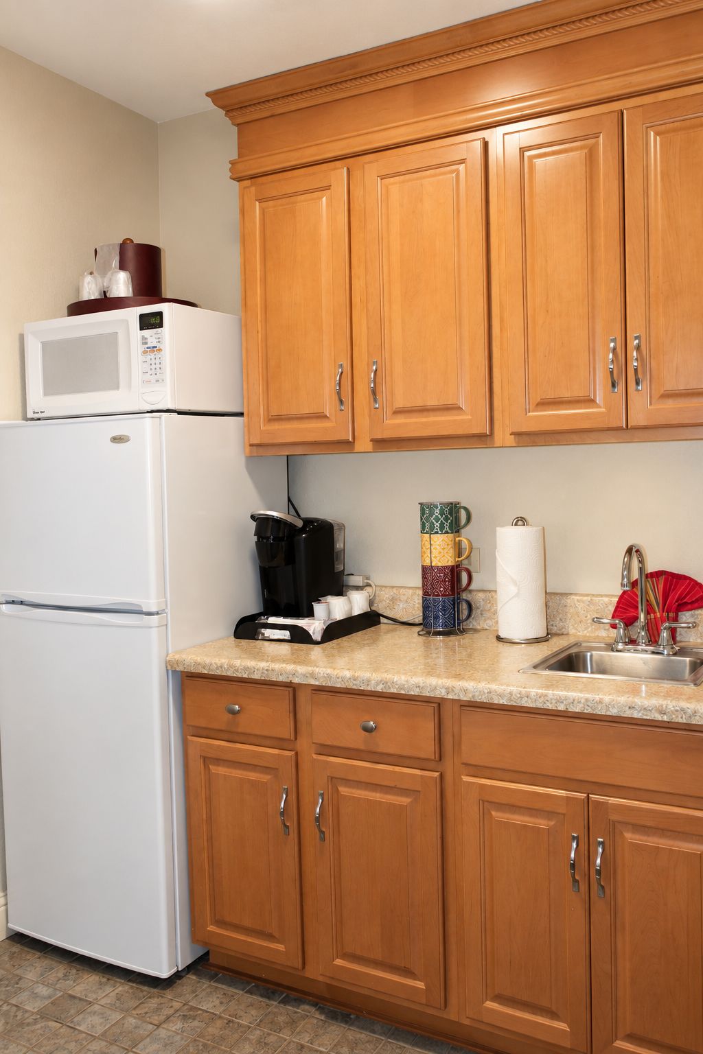 A kitchen with wooden cabinets, a white refrigerator, a microwave, a coffee maker, a stack of mugs, and a sink.