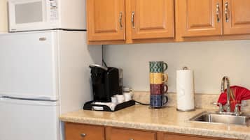 A kitchen with wooden cabinets, a white refrigerator, a microwave, a coffee maker, a stack of mugs, and a sink.