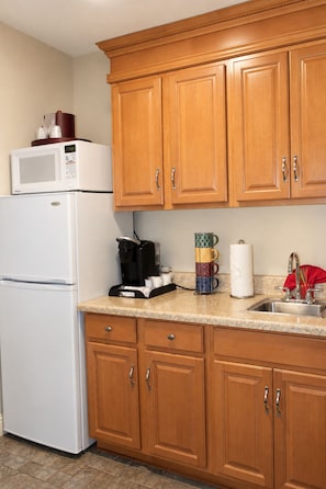 A kitchen with wooden cabinets, a white refrigerator, a microwave, a coffee maker, a stack of mugs, and a sink.