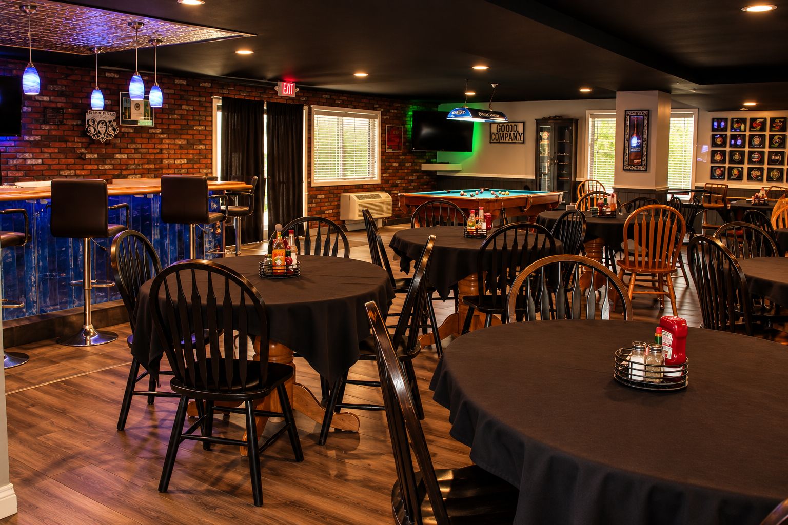 Zona de bar con mesas y sillas, una mesa de billar y una pared de ladrillo con un televisor.