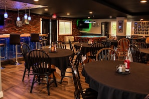 Zona de bar con mesas y sillas, una mesa de billar y una pared de ladrillo con un televisor.