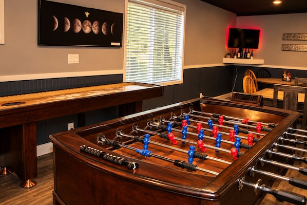 A room with a foosball table, a billiards table, and a wall-mounted TV.