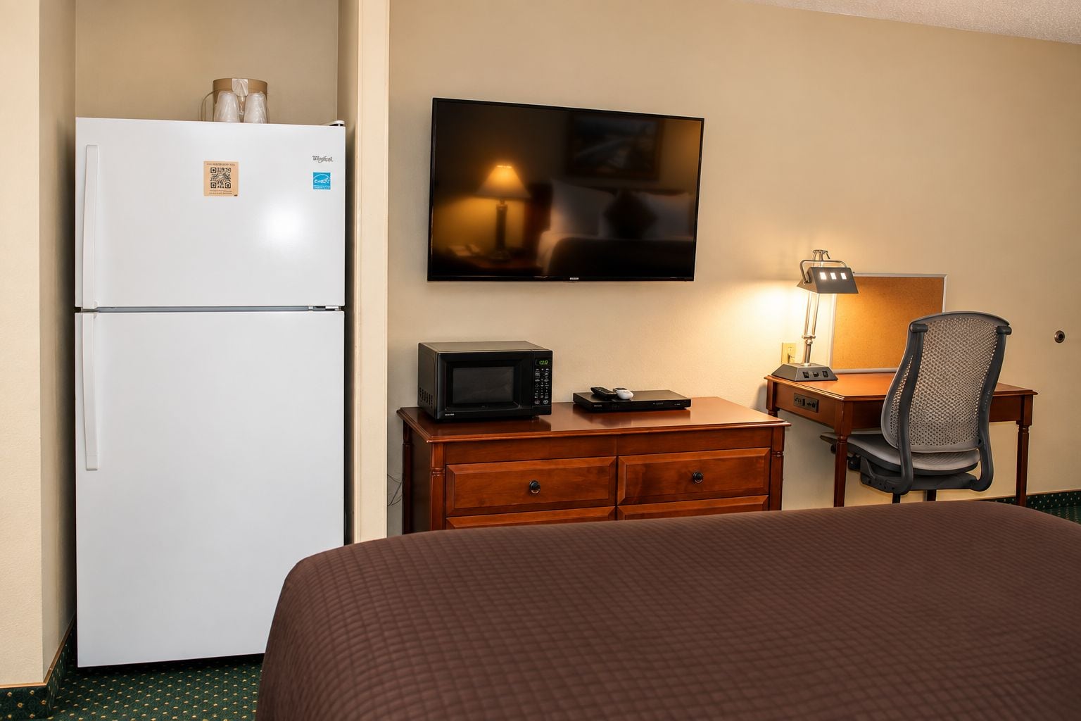 A hotel room with a bed, a desk with a computer, a microwave, a refrigerator, and a flat-screen TV mounted on the wall.
