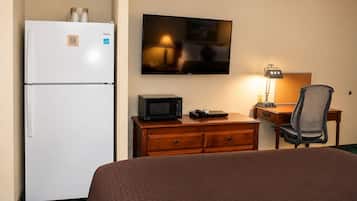 A hotel room with a bed, a desk with a computer, a microwave, a refrigerator, and a flat-screen TV mounted on the wall.