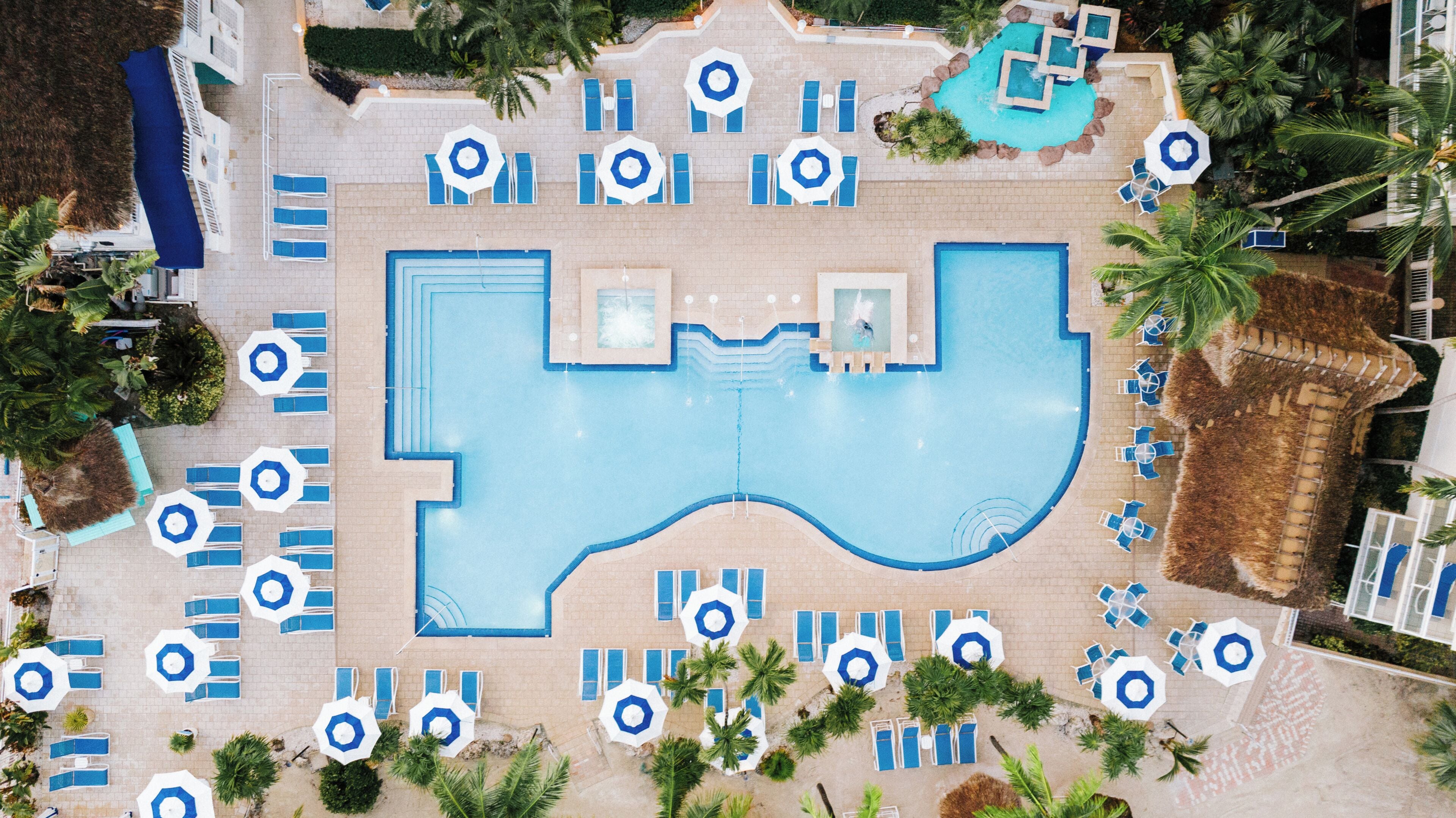 An aerial view of a resort with a large central pool, multiple smaller pools, and numerous sun loungers and umbrellas.