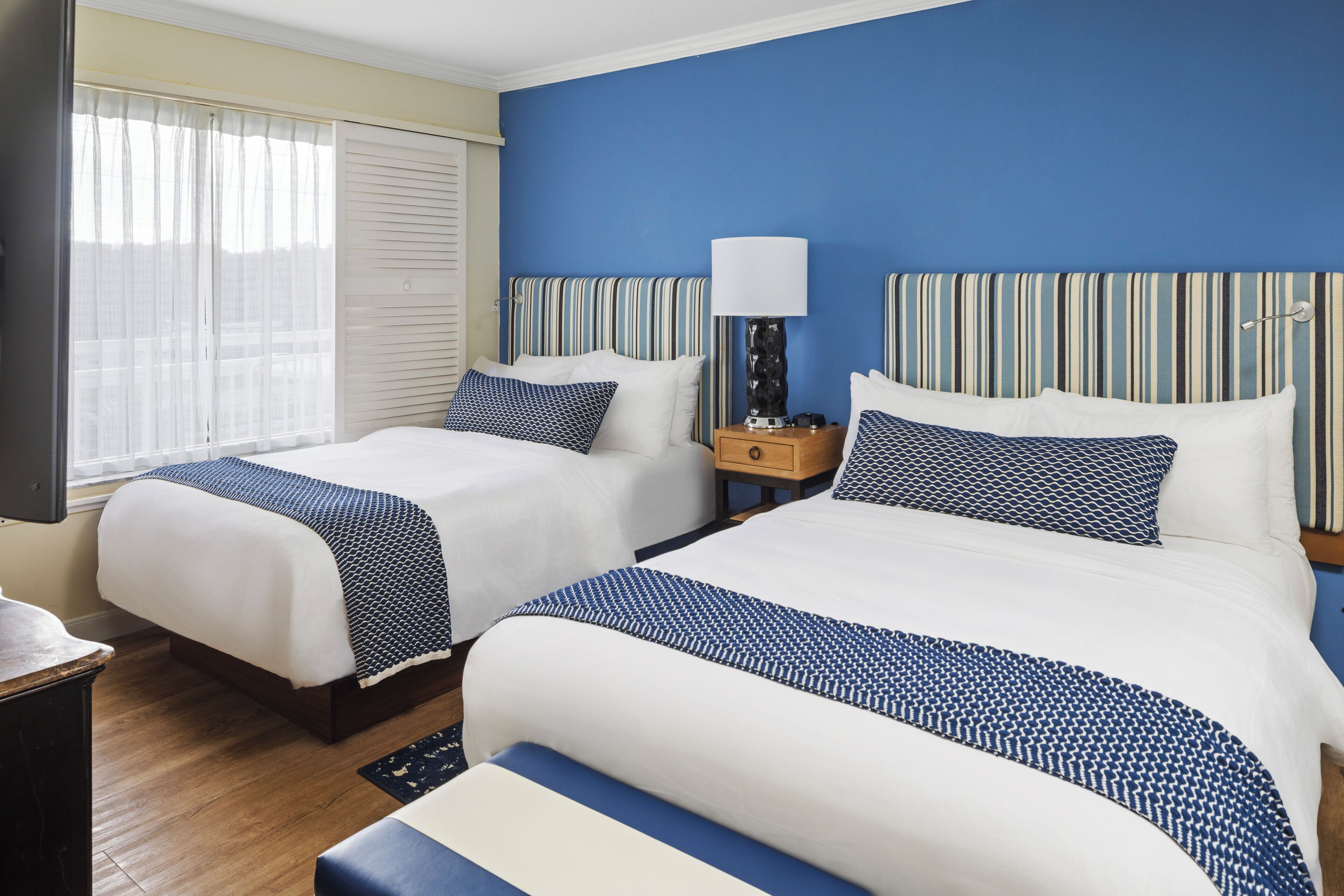 A hotel room with two beds, a TV, a nightstand, and a window with blinds.
