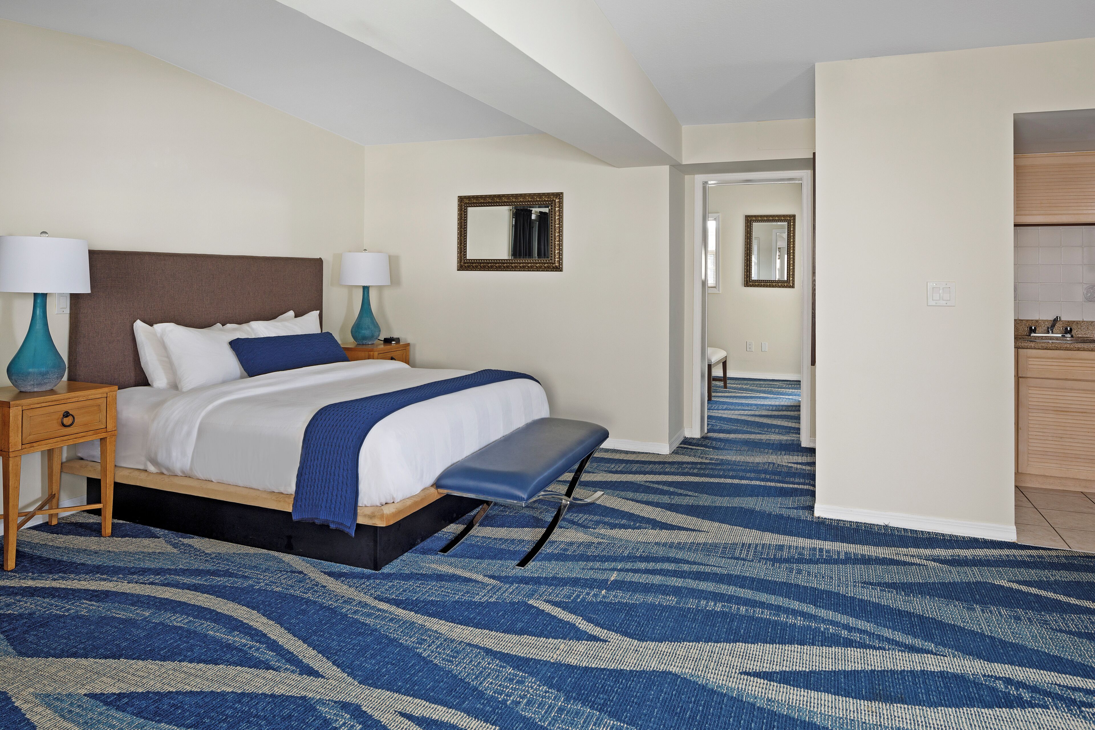 A hotel room with a bed, bedside tables, a bench, a mirror, and a view into the bathroom.