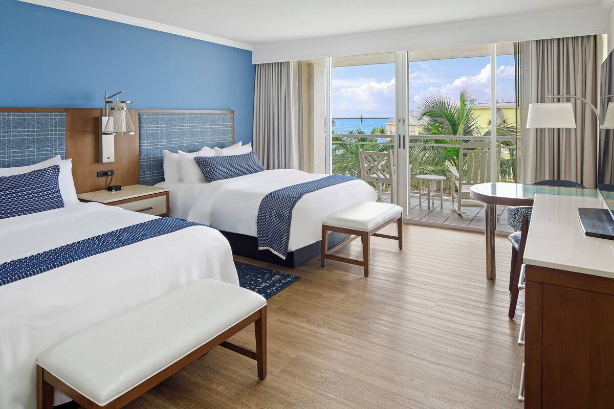 Ein Hotelzimmer mit zwei Betten, einem Schreibtisch, einem Stuhl und einem Balkon mit Meerblick.