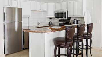 A kitchen with white cabinets, stainless steel appliances, a granite countertop, and three leather bar stools.