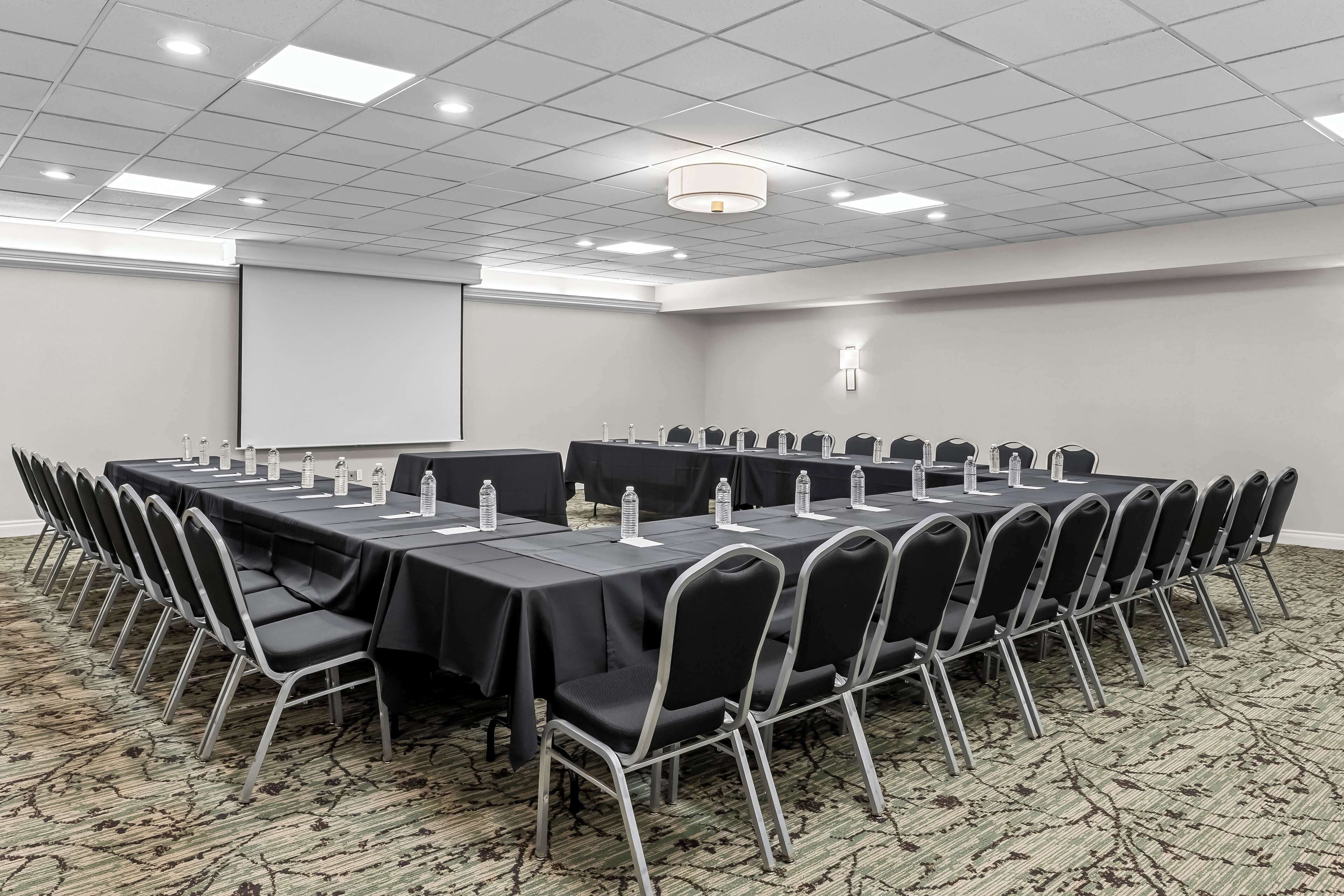 Un salón de conferencias con una larga mesa, sillas, botellas de agua y una pantalla para proyector.