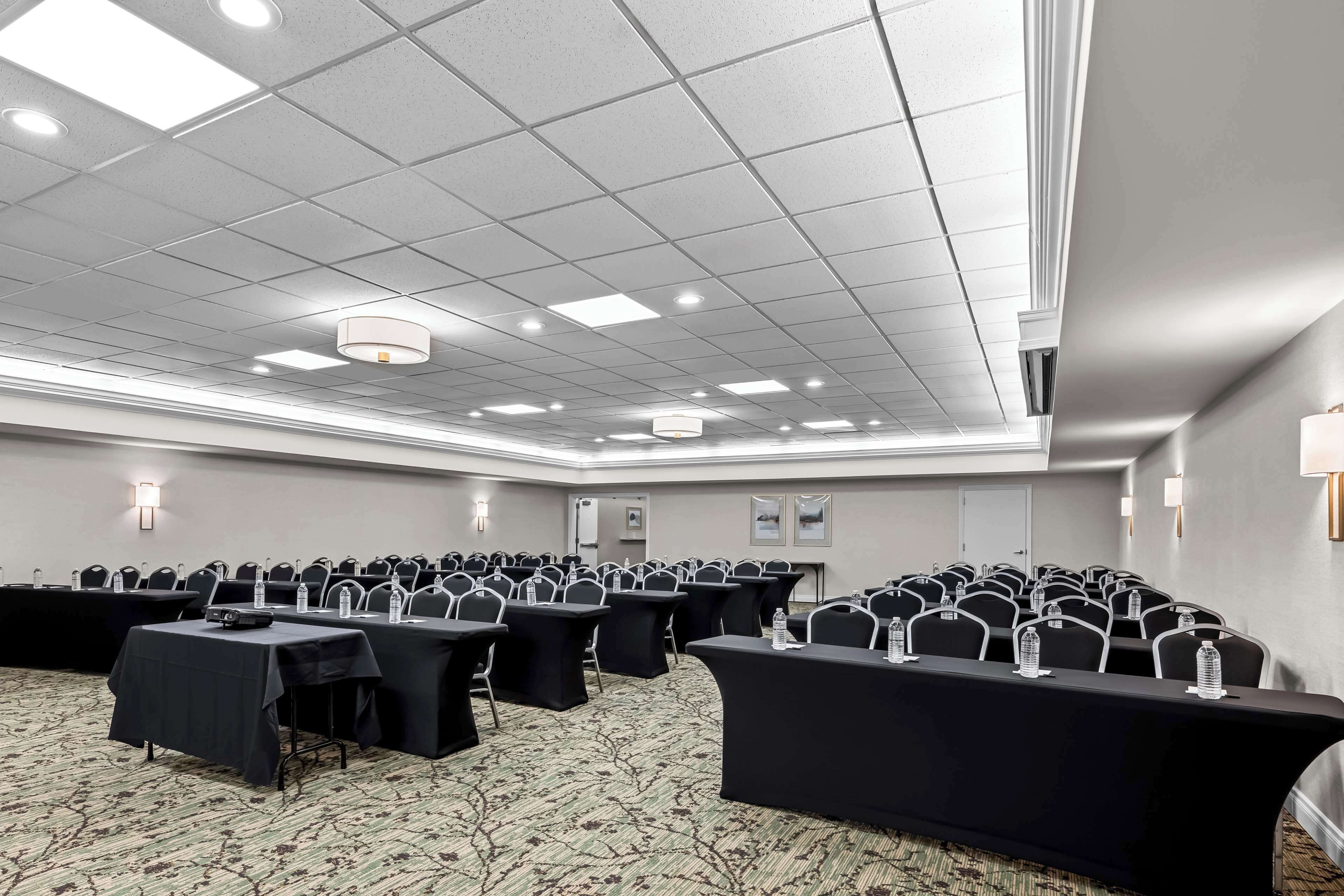 A conference room with rows of tables and chairs, ceiling lights, and a carpeted floor.