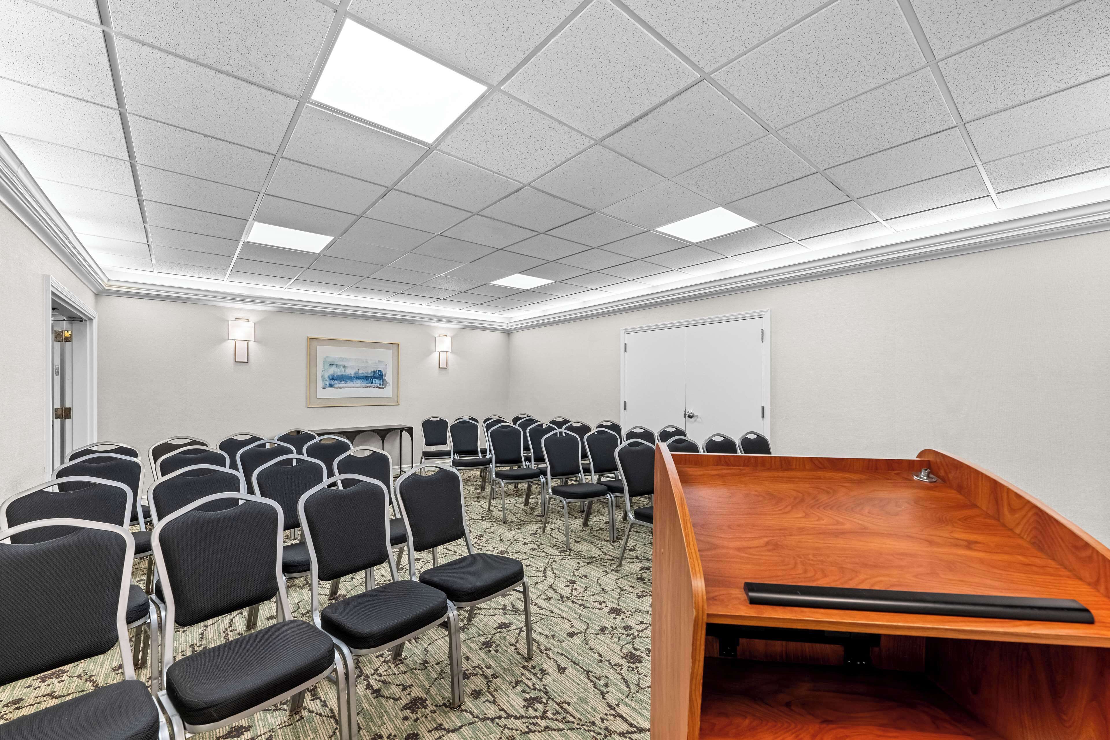A conference room with rows of chairs, a wooden presentation table, and a white ceiling with fluorescent lights.