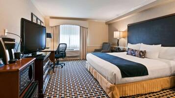 Quarto de hotel com uma cama grande, uma escrivaninha com televisão, uma cadeira e uma janela com cortinas.