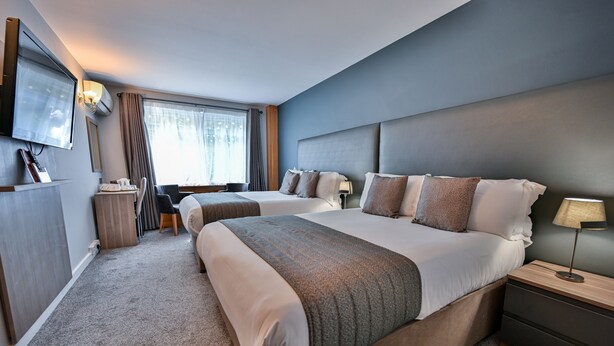 A hotel room with two beds, a TV mounted on the wall, a desk with a chair, and a window with curtains. - Kensington Court Hotel (London)