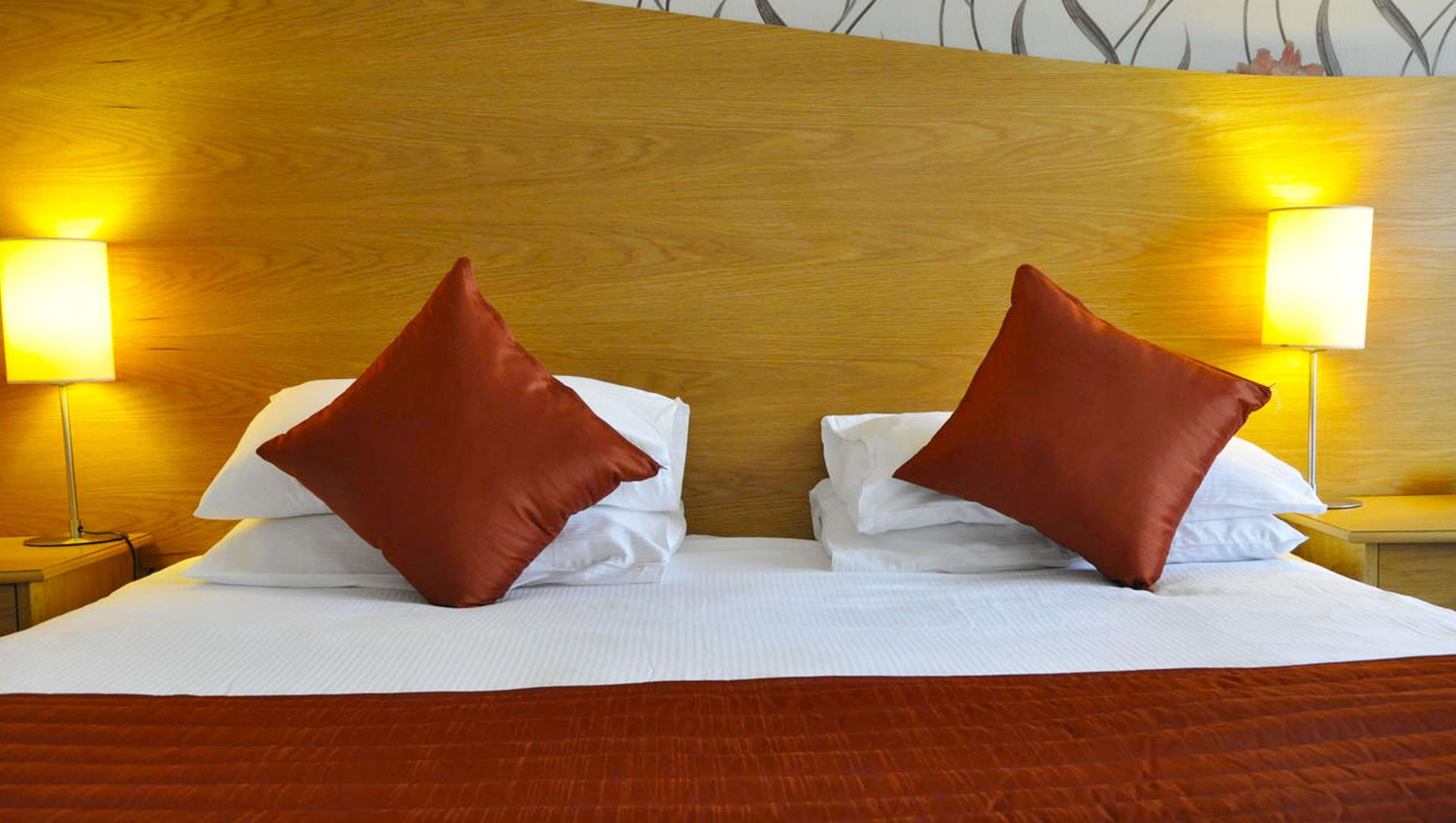 A neatly made bed with two red pillows and white linens, flanked by two bedside lamps on wooden nightstands.