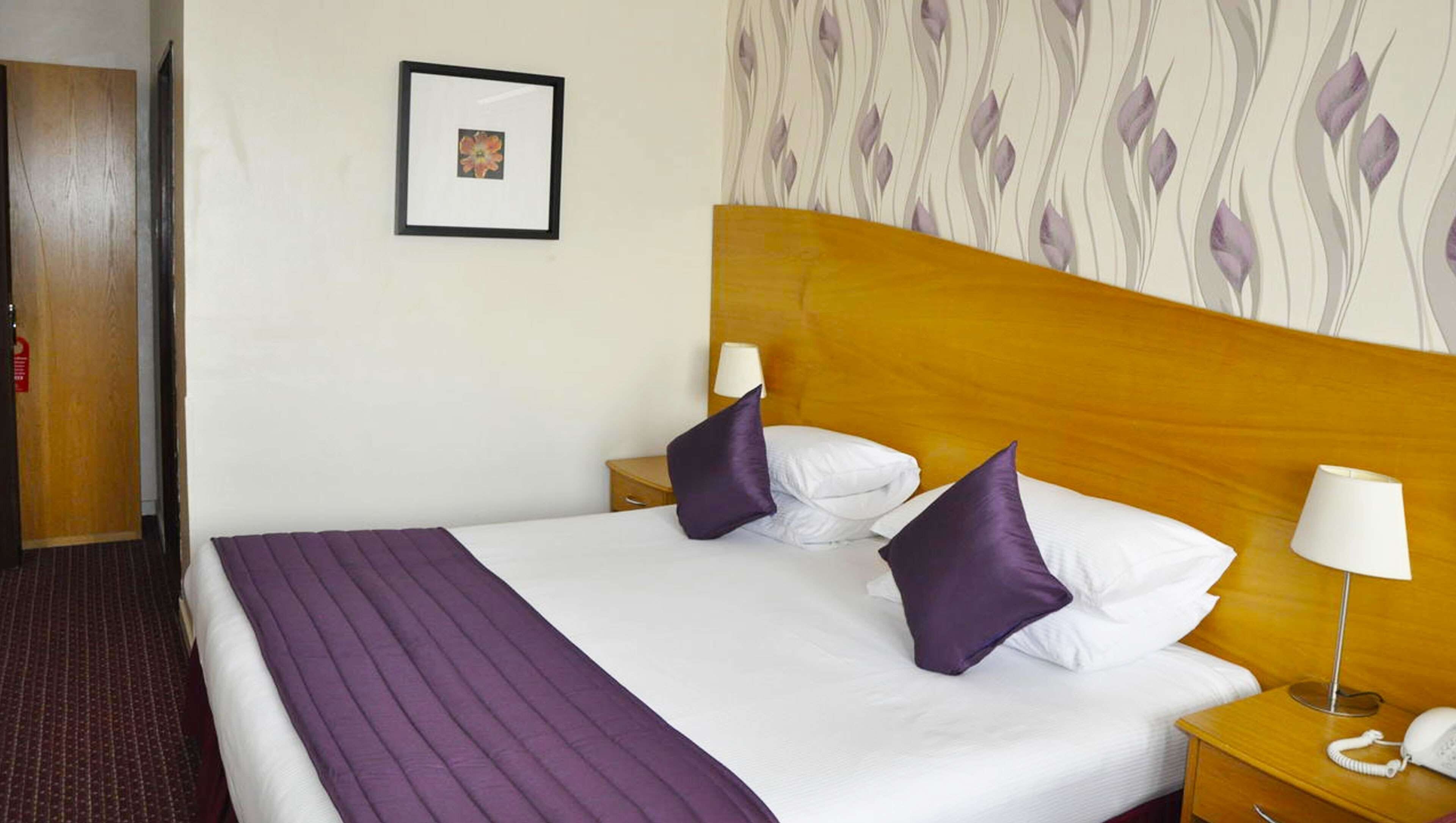 A hotel room with a large bed, two nightstands, a wooden headboard, a framed picture on the wall, and a patterned wallpaper.