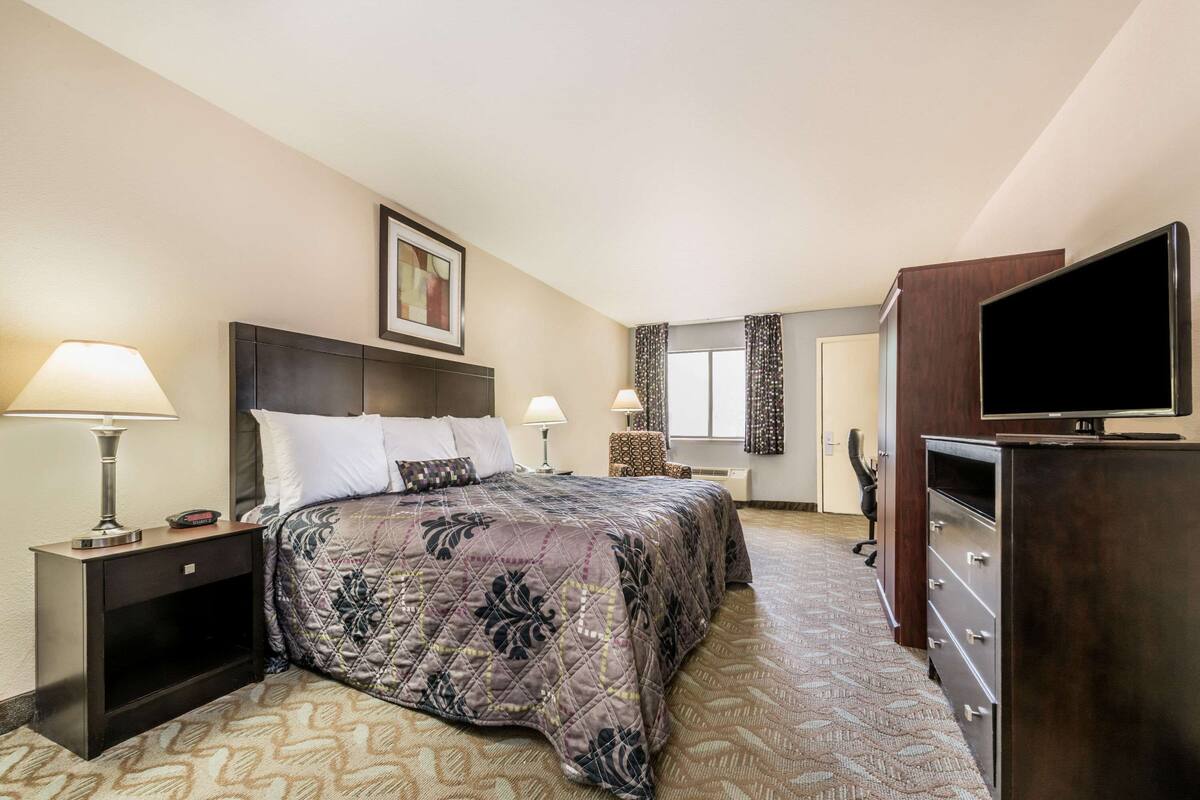 A hotel room with a bed, two bedside tables with lamps, a dresser with a TV, and a window with curtains.