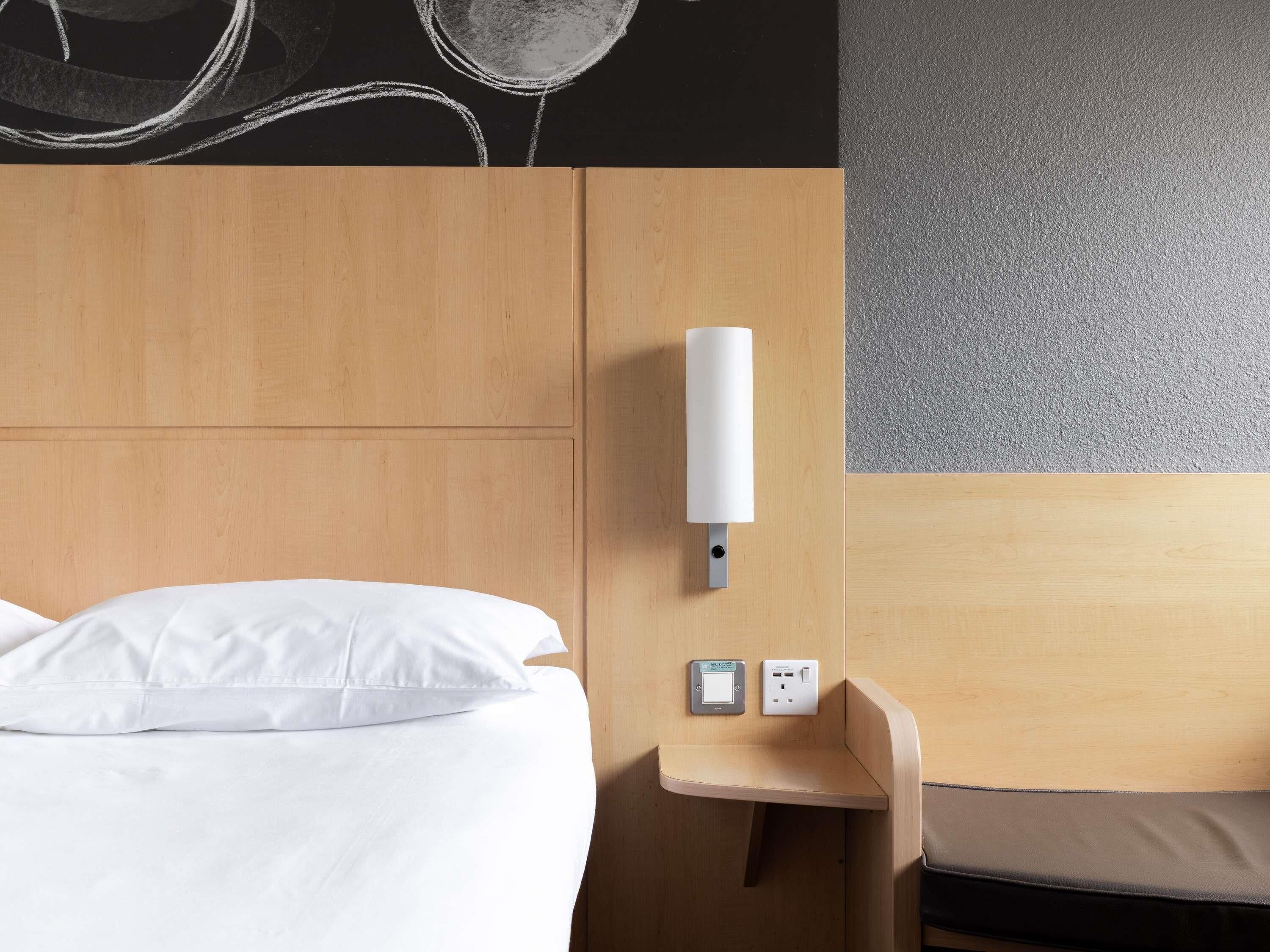 Habitación de hotel con cama, mesita de noche, luz empotrada en la pared y toma de corriente.