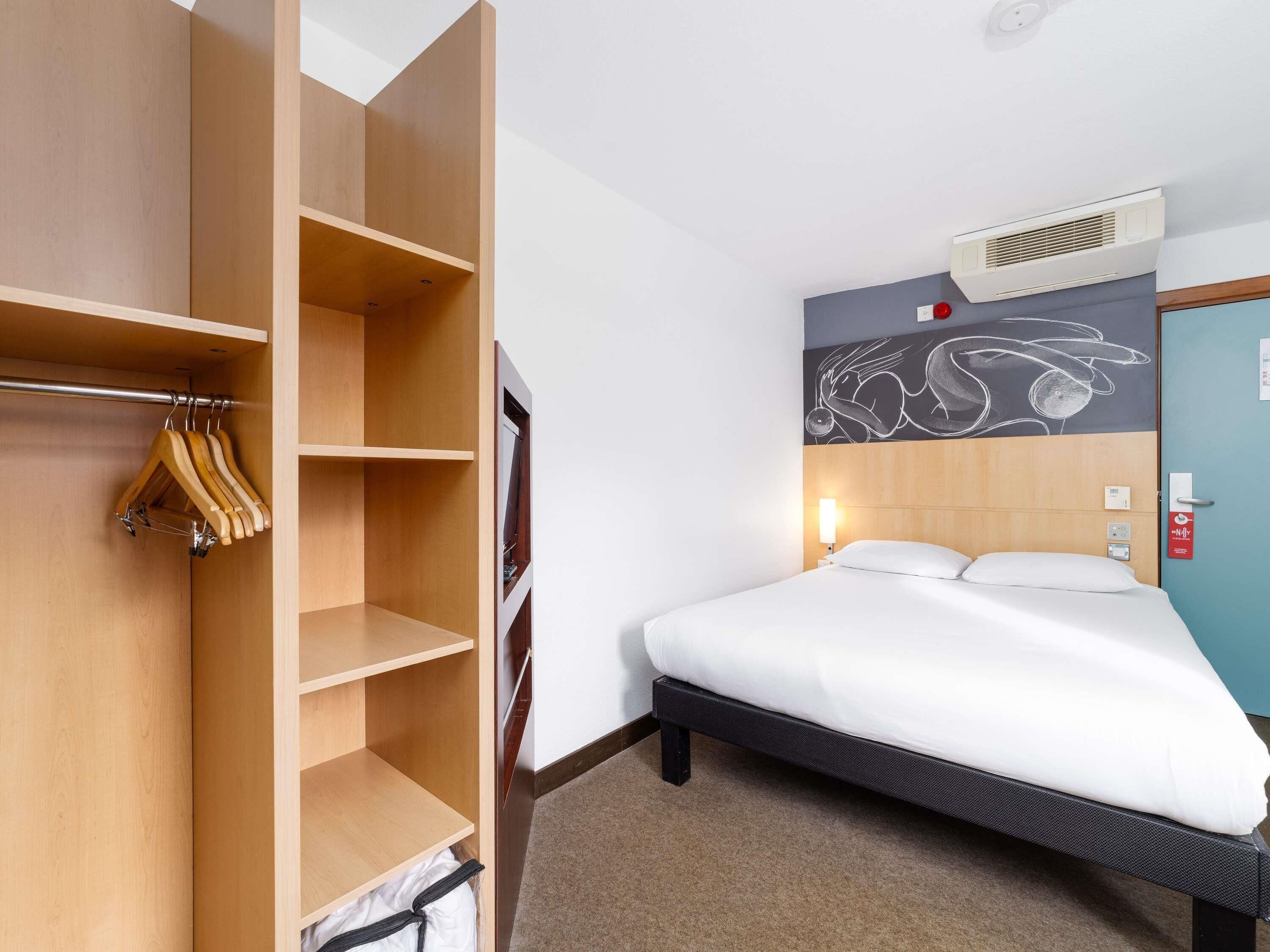 Una habitación de hotel con una cama, un armario con ropa colgada, un aire acondicionado instalado en la pared y una puerta con un letrero rojo.