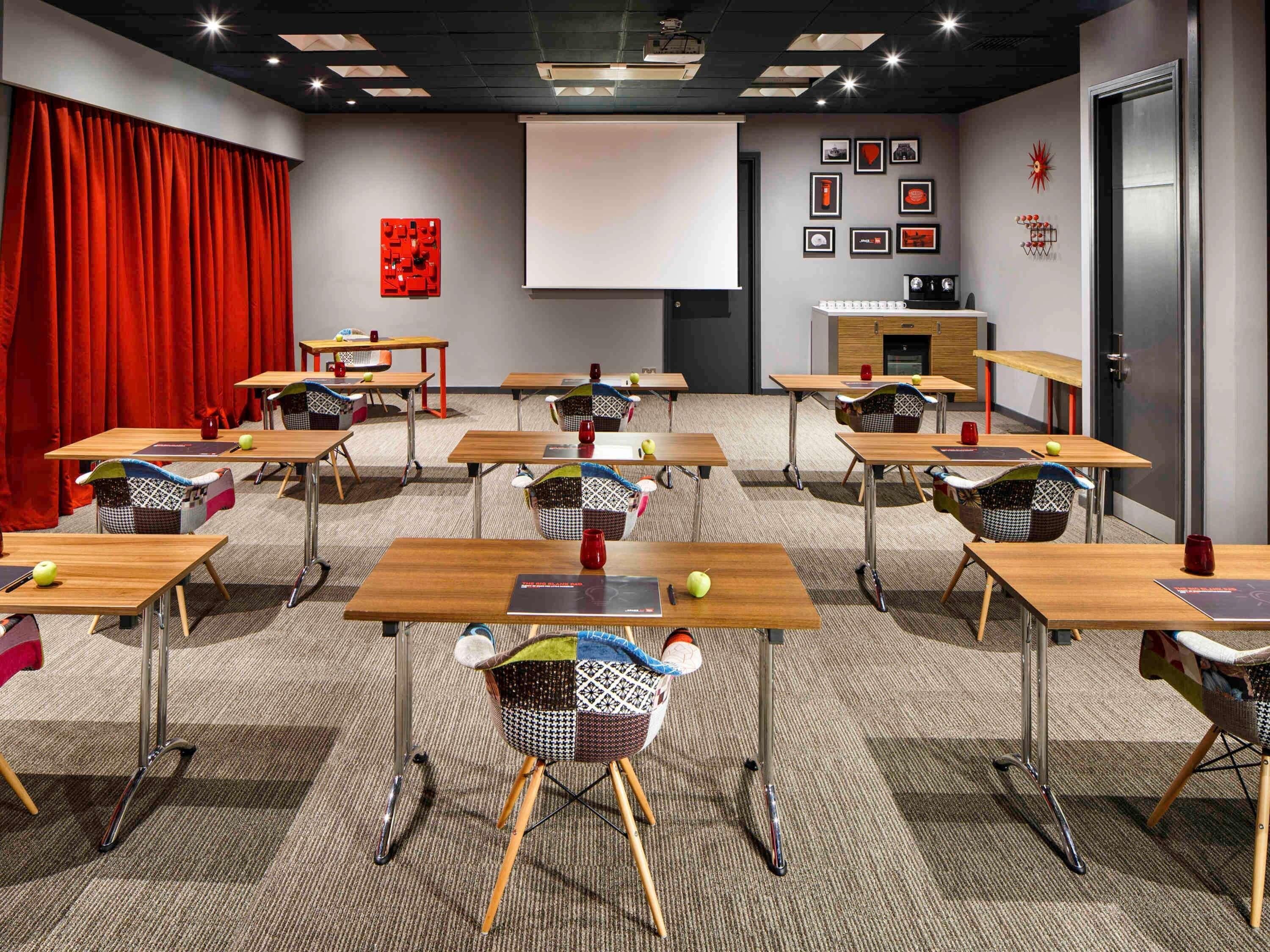 Un salón de conferencias con mesas, sillas, una pantalla de proyector y una cortina roja.