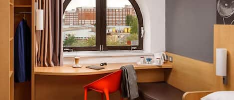 A hotel room with a bed, a desk with a chair, a window with curtains, a view of buildings, and a wall-mounted fan.