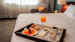 Una cama bien hecha con una bandeja de desayuno que incluye jugo de naranja, croissants y bollería, en una habitación de hotel con una silla y una lámpara al fondo.