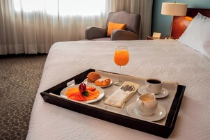 Una cama bien hecha con una bandeja de desayuno que incluye jugo de naranja, croissants y bollería, en una habitación de hotel con una silla y una lámpara al fondo.