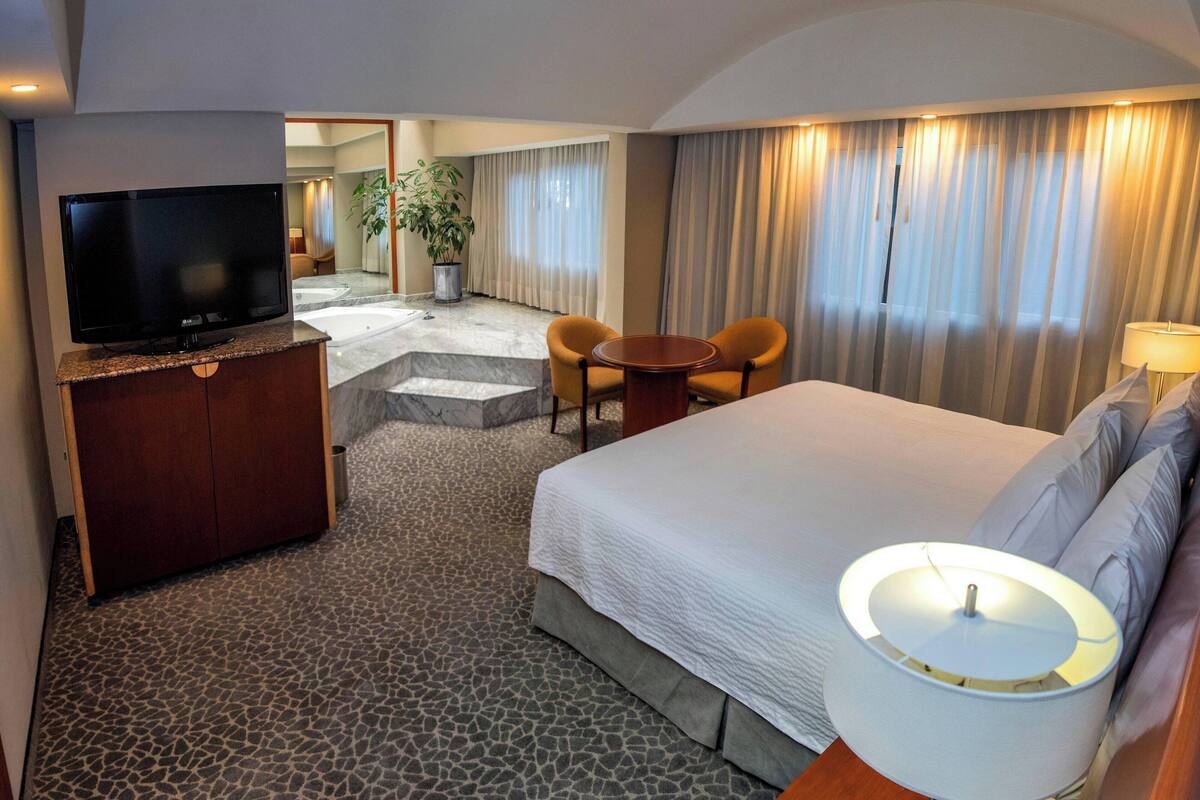 Una habitación de hotel con una cama, televisión, escritorio con silla, una mesita y una lámpara.