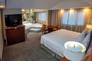 Una habitación de hotel con una cama, televisión, escritorio con silla, una mesita y una lámpara.