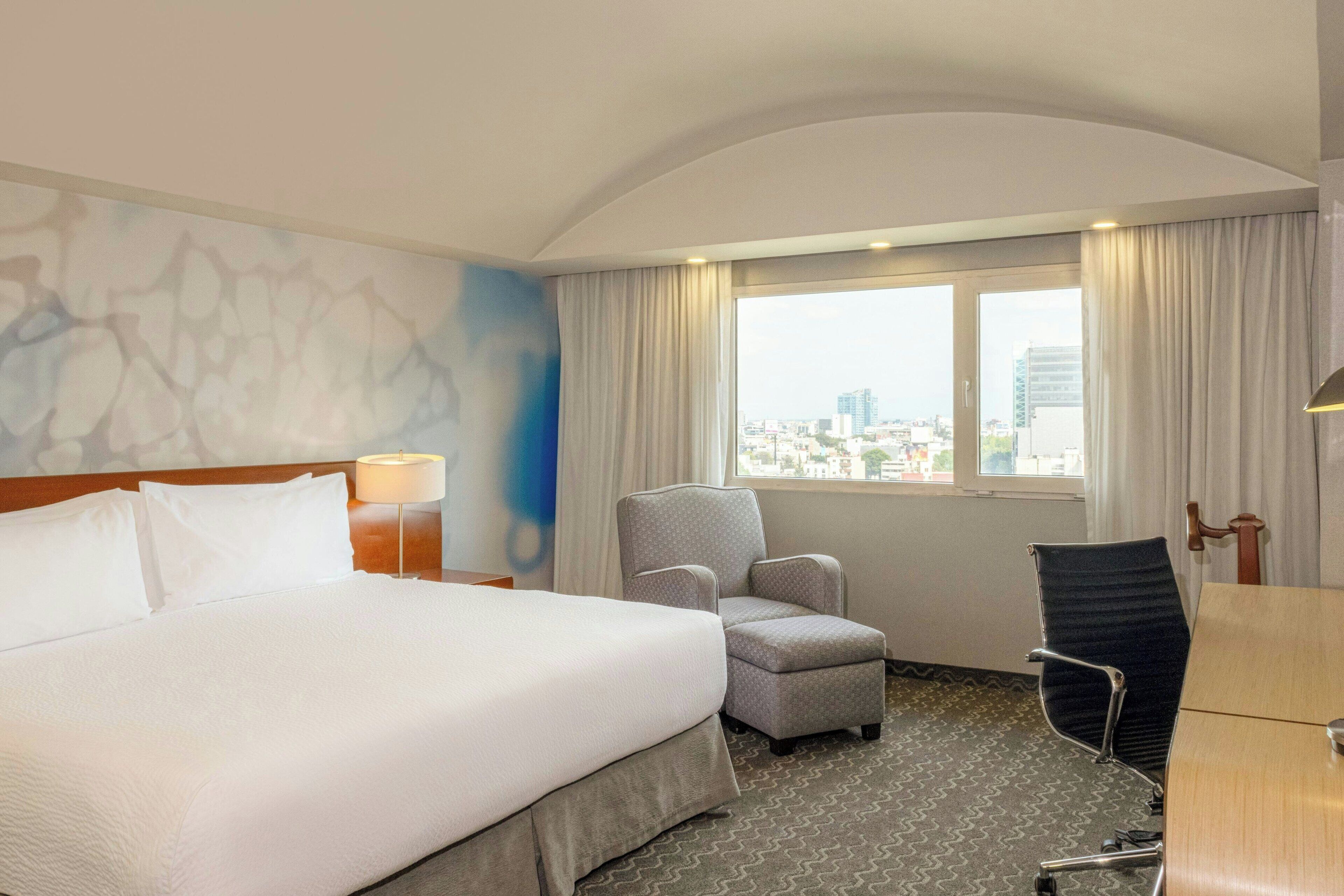 Une chambre d’hôtel avec un grand lit, deux fauteuils, un bureau et une vue sur la ville par la fenêtre.