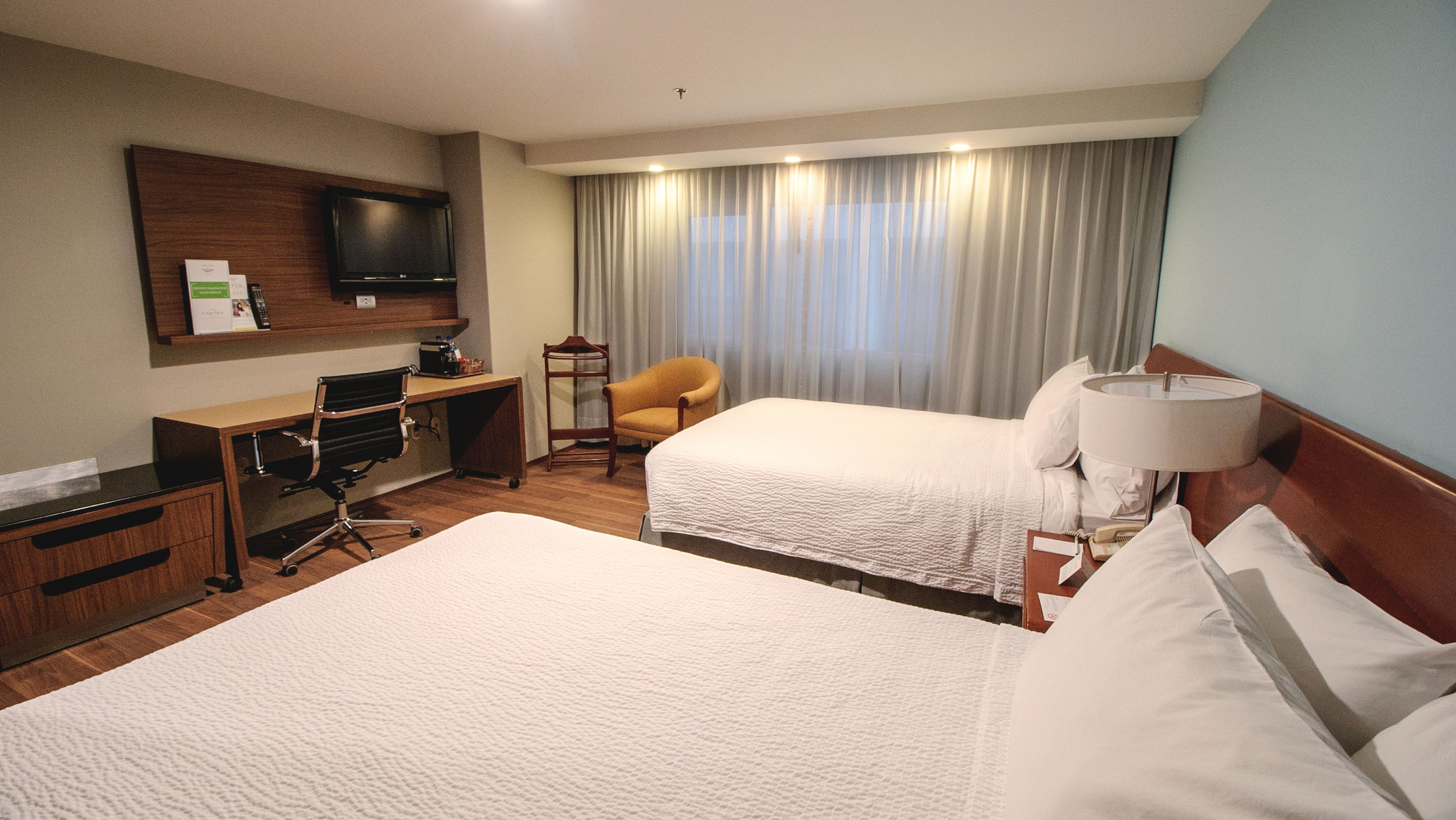 Une chambre d’hôtel avec deux lits, un bureau avec un ordinateur, une chaise, une télévision et une fenêtre avec des rideaux.