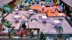 Un patio interior amplio con varias zonas de estar, incluyendo mesas redondas y sofás, rodeado de plantas en macetas e iluminación decorativa.
