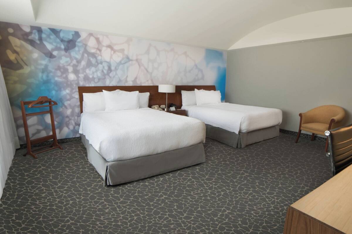 Una habitación de hotel con dos camas, una silla de madera, un escritorio y un papel tapiz estampado.
