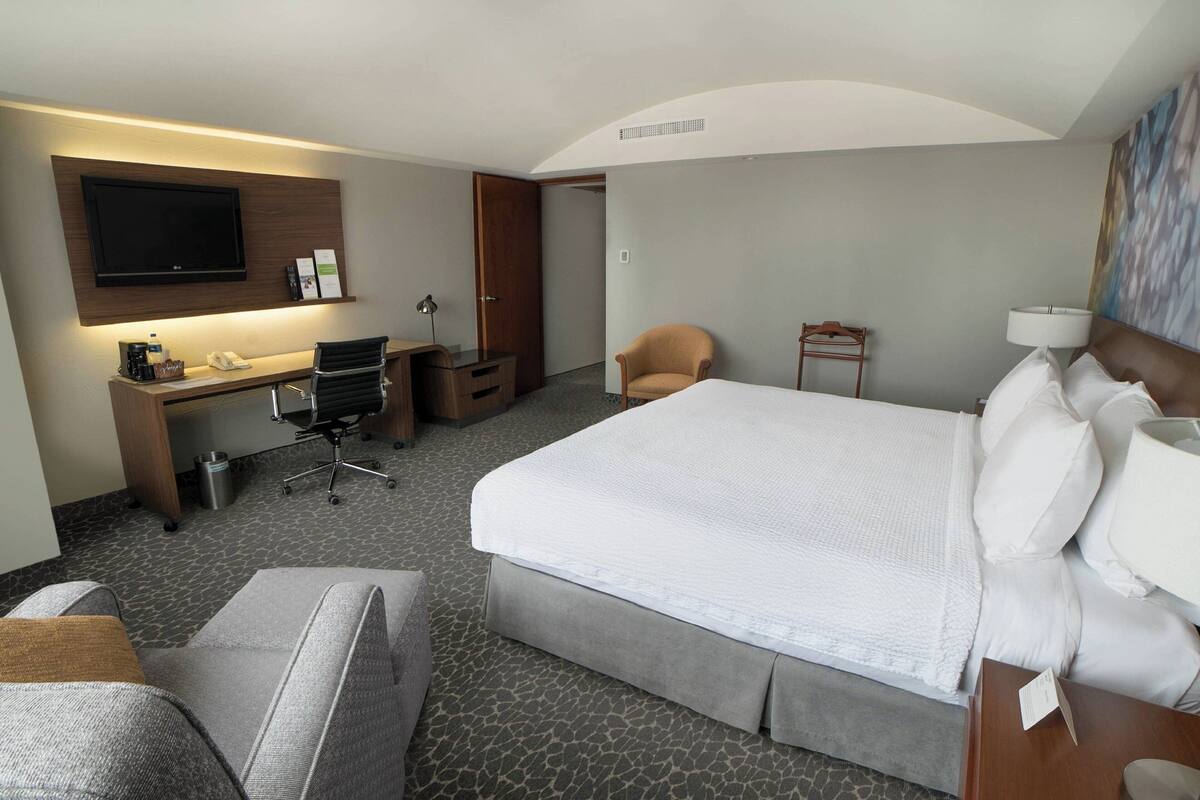 Habitación de hotel con una cama grande, un escritorio con computadora, una silla, un televisor montado en la pared y un cuadro en la pared.