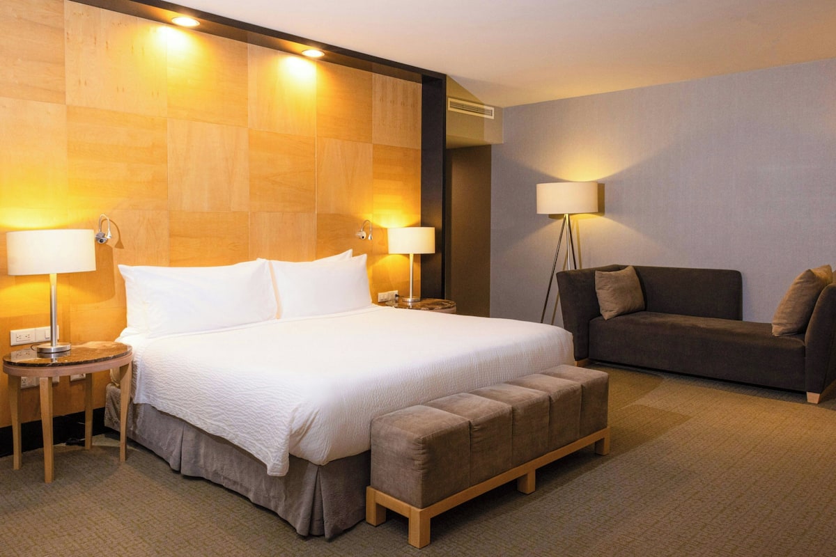 Una habitación de hotel con una cama grande, dos mesitas de noche con lámparas, un sofá y un reposapiés.