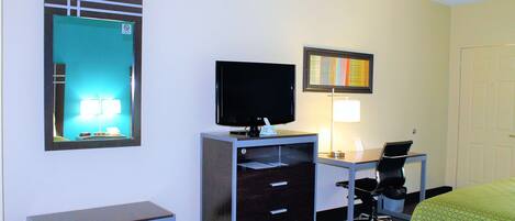 A hotel room with a desk, a television, a chair, a bed, and a mirror.