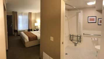 A hotel room with a bed, a bedside table with a lamp, a window with curtains, and a bathroom with a shower, toilet, and towel rack.