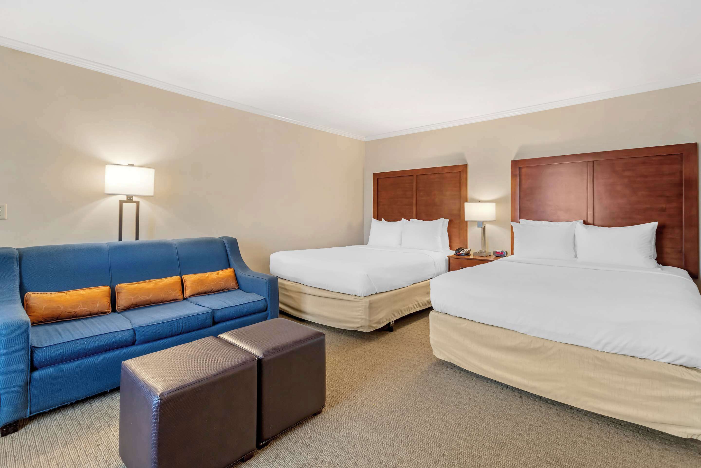 A hotel room with a blue sofa, two single beds, a brown ottoman, and a nightstand with a lamp.