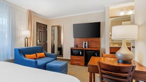 Habitación de hotel con una cama grande, un sofá azul con cojines naranja, una mesa de comedor de madera, un televisor montado en la pared y una pequeña cocina americana con microondas y cafetera.