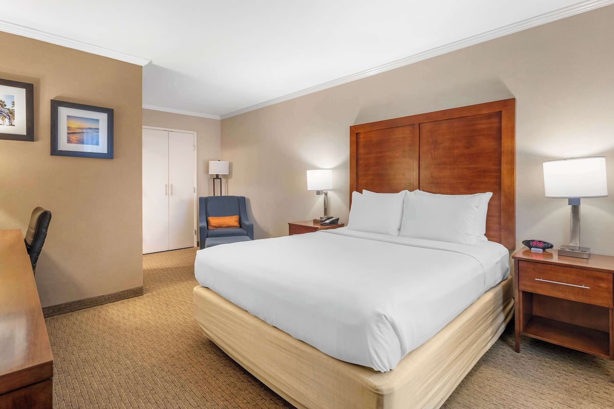 A hotel room with a large bed, a desk, a chair, and a nightstand with a lamp.
