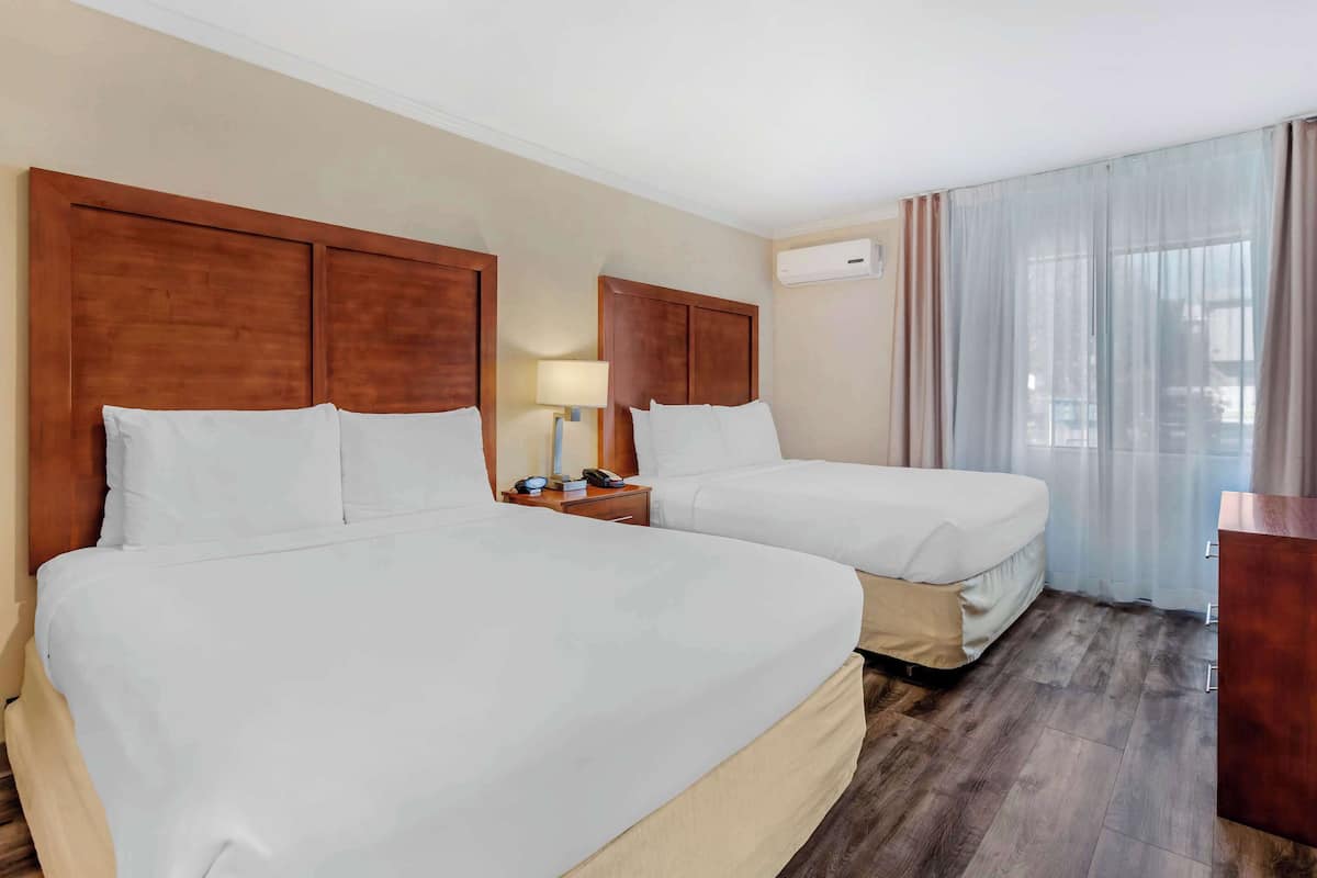 A hotel room with two beds, a nightstand with a lamp, an air conditioning unit, and a view of the city through sheer curtains.