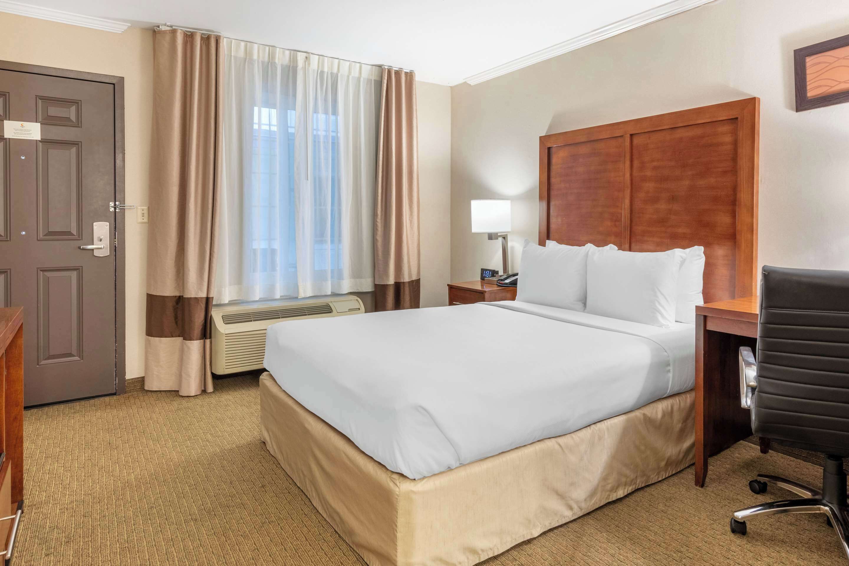 Habitación de hotel con una cama grande, un escritorio con silla, una mesita de noche con lámpara y una ventana con cortinas.