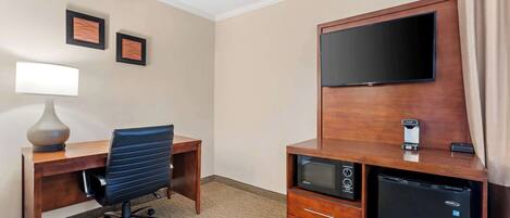 A hotel room with a wooden desk, a black office chair, a flat-screen TV mounted on the wall, a small refrigerator, and a microwave.