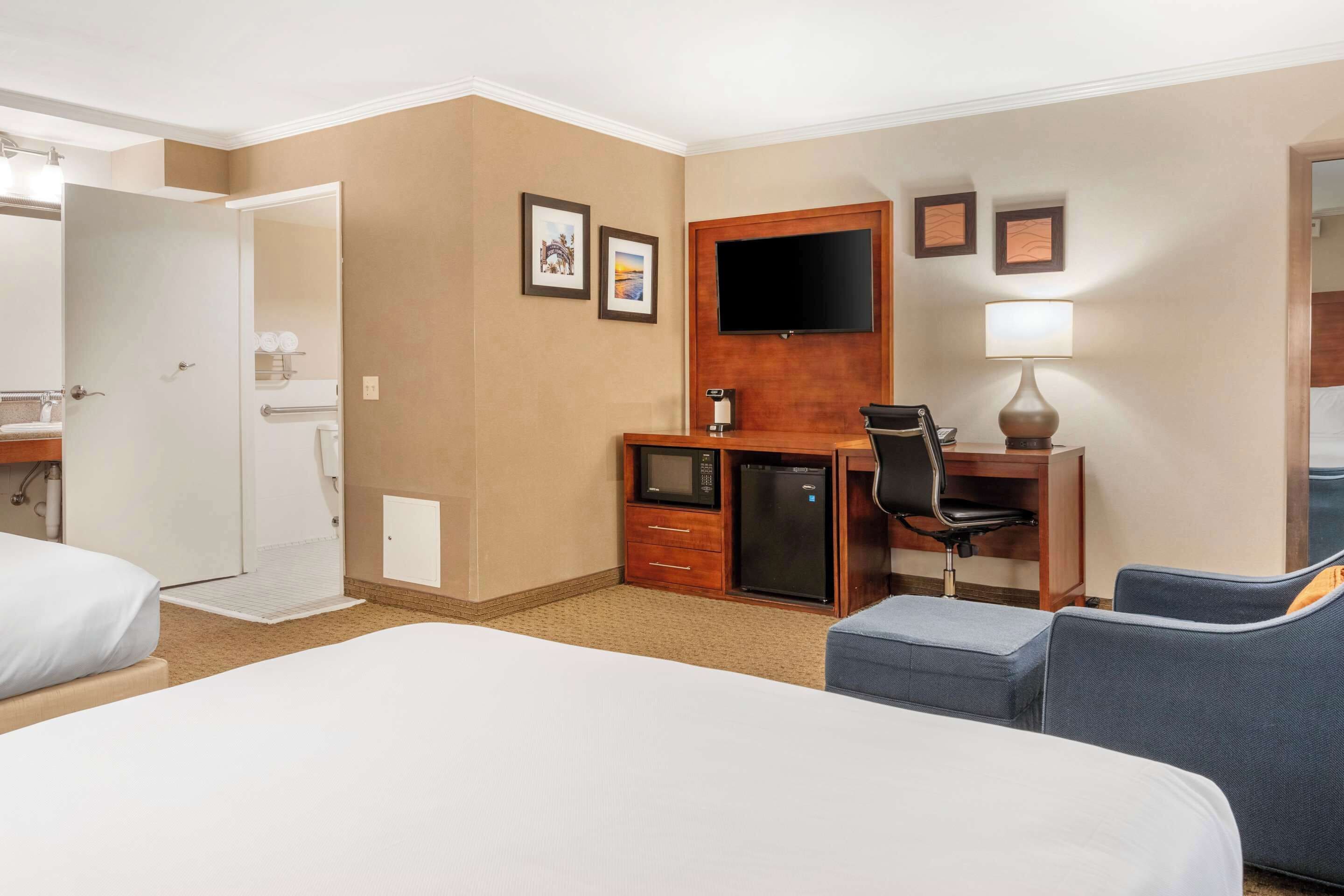 A hotel room with two beds, a desk with a computer, a TV, and a small kitchenette.