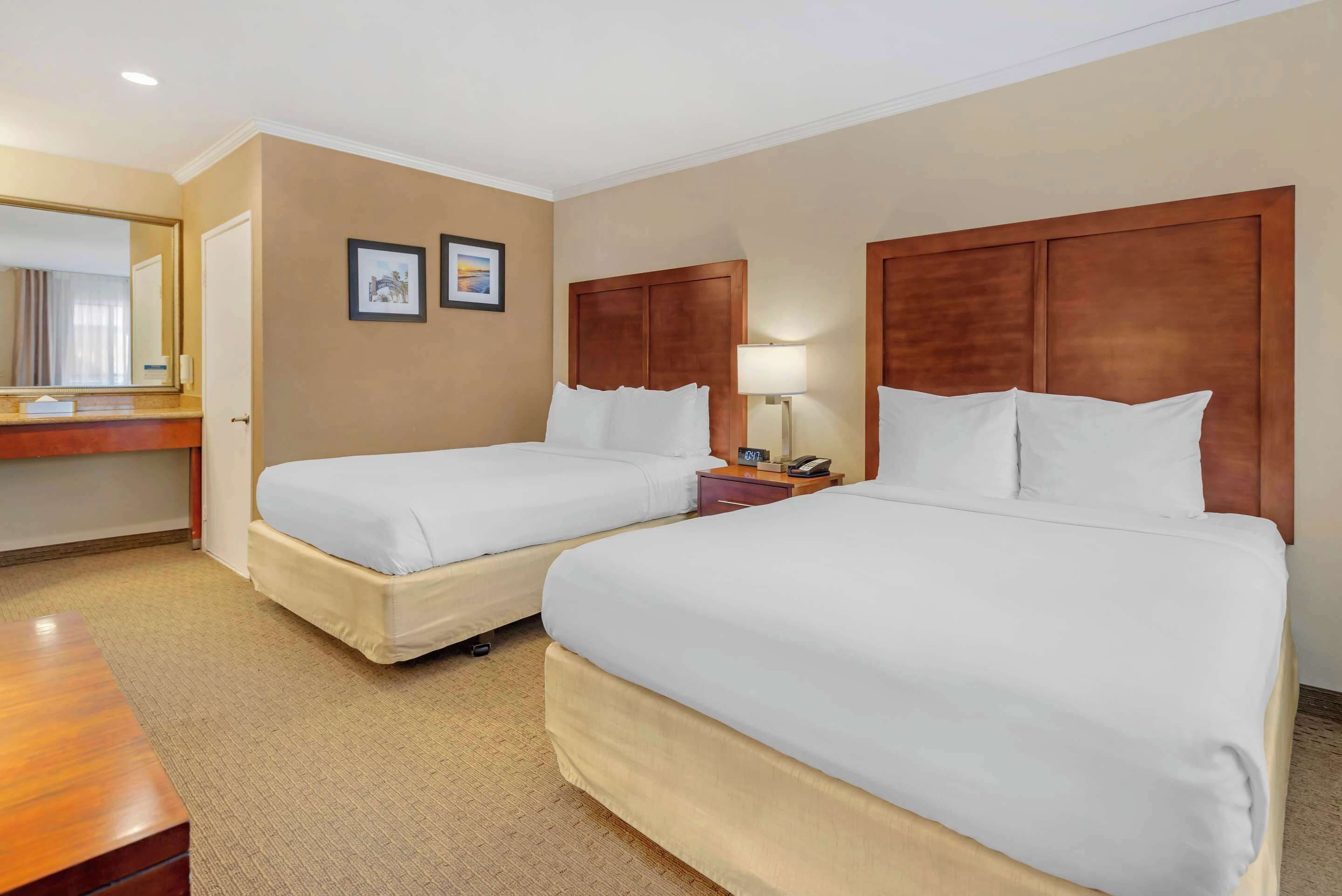 Quarto de hotel com duas camas, cabeceira de madeira, mesa de cabeceira com abajur e um espelho grande.