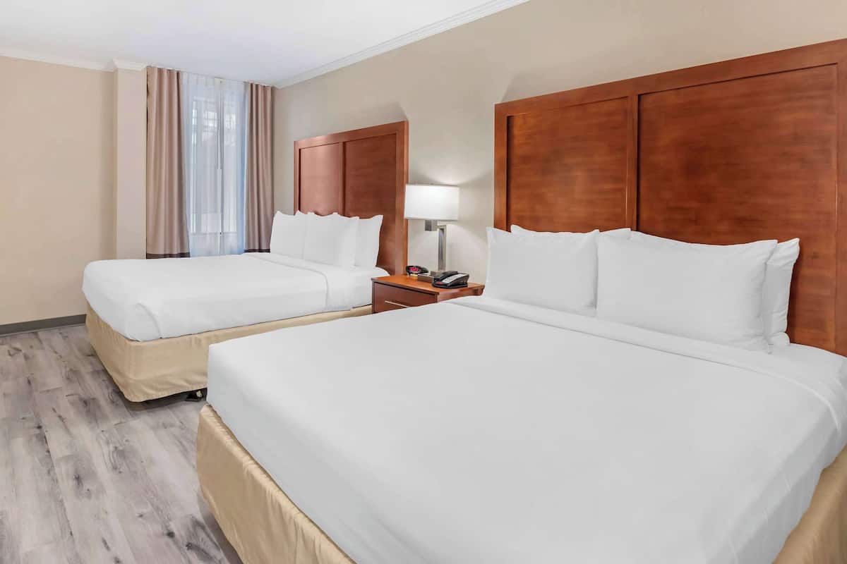 A hotel room with two beds, wooden headboards, a nightstand with a lamp, and a window with sheer curtains.